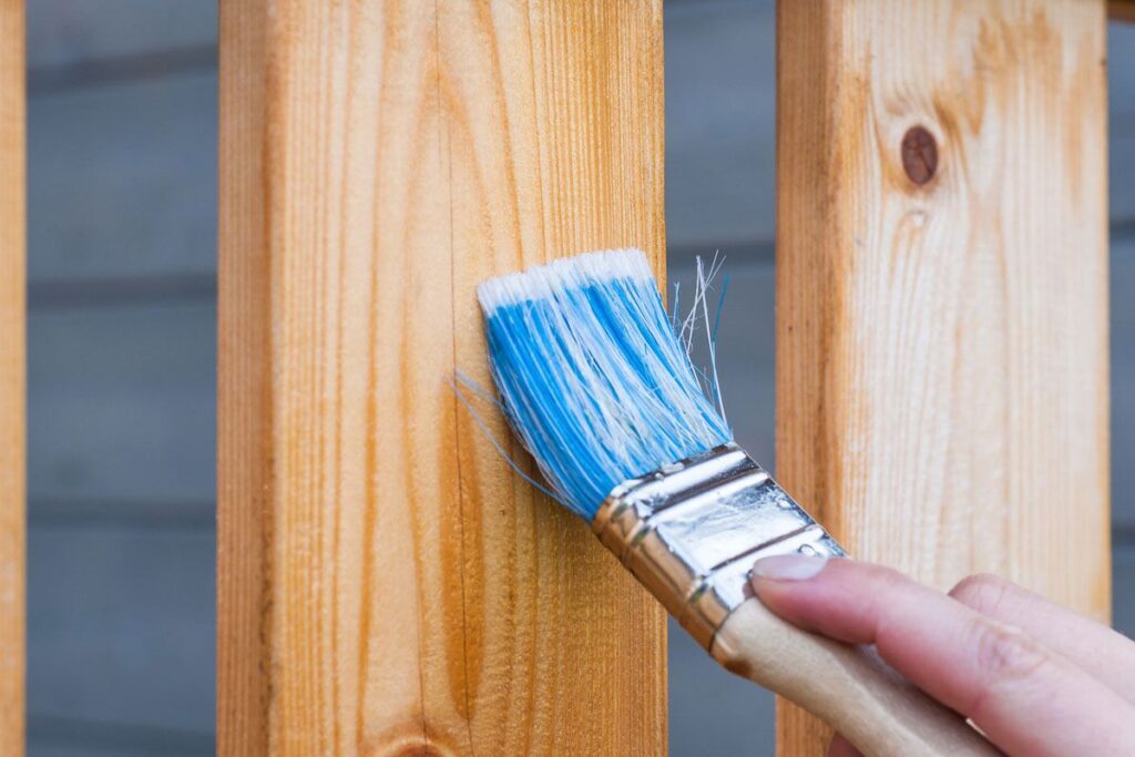 A person holding a brush over the top of a wooden fence.